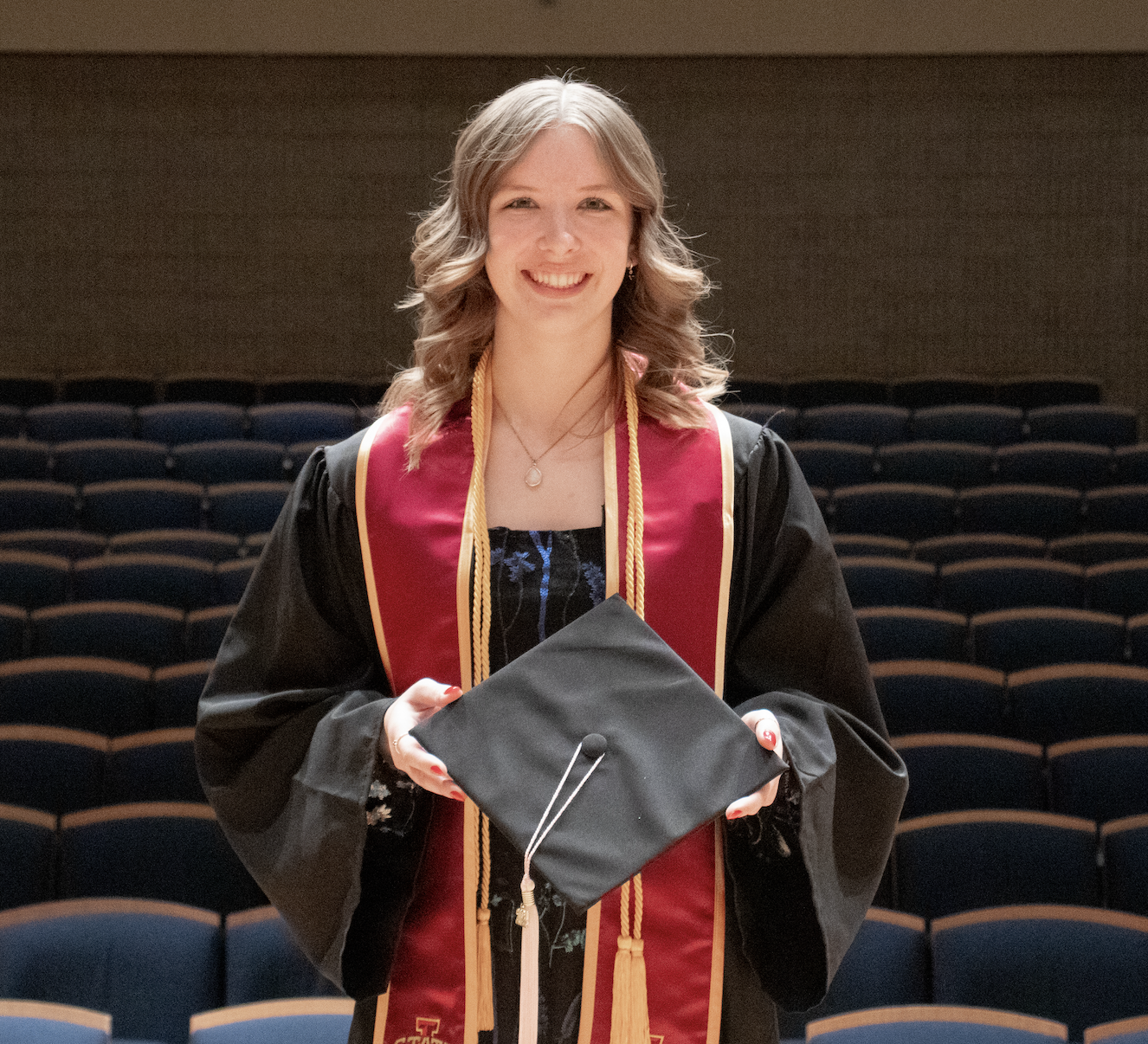 Jaslyn Riherd stands smiling in an auditorium. She is wearing a black graduation gown with red and gold honor cords and a matching red stole that reads "STATE" with an emblem. She holds her black graduation cap in front of her with both hands. The auditorium behind her is filled with rows of empty blue seats, and the lighting softly highlights her and the setting.